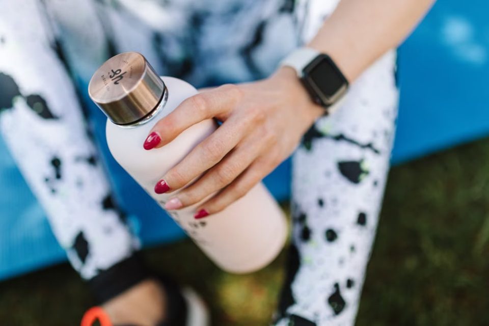 Crop unrecognizable female wearing sports leggings smart watch and holding bottle with water while relaxing after training on mat outside